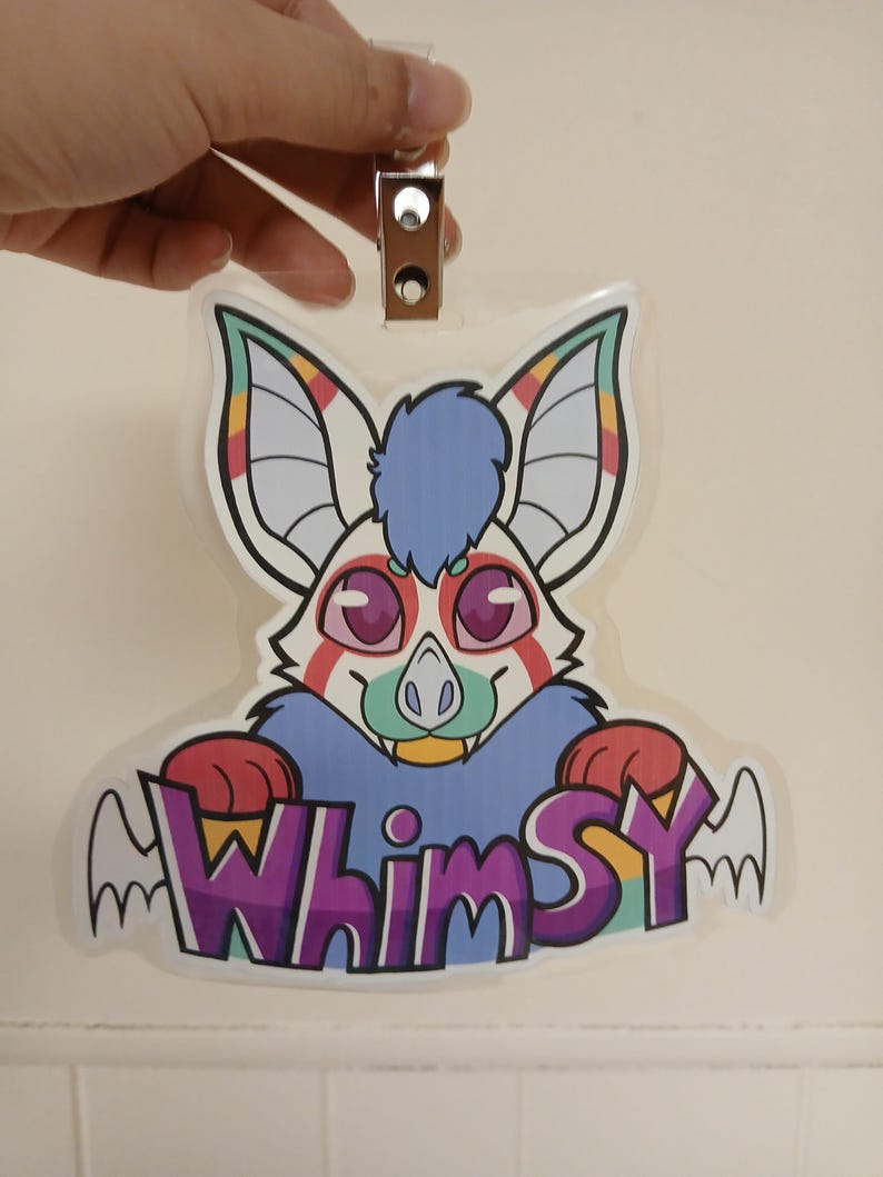 May include: A clear, plastic badge featuring a cartoon bat with large ears and a blue tuft of fur. The bat has pink eyes, a green nose, and a smiling mouth. The word "Whimsy" is written in purple and yellow letters with wing accents.