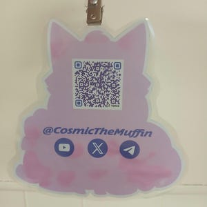May include: A light purple badge in the shape of a cat with a QR code and social media icons. The text "@CosmicTheMuffin" is displayed below the QR code. The badge is attached to a lanyard with a metal clip.