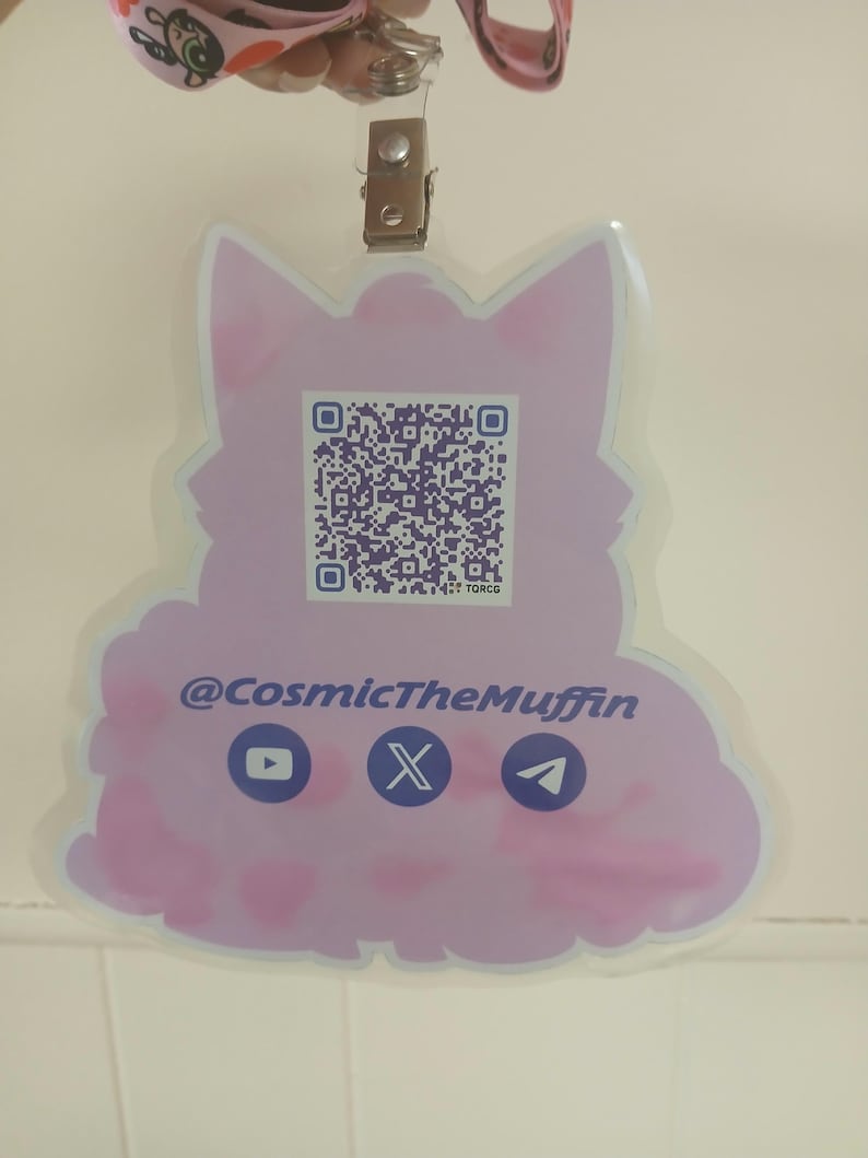 May include: A light purple badge in the shape of a cat with a QR code and social media icons. The text "@CosmicTheMuffin" is displayed below the QR code. The badge is attached to a lanyard with a metal clip.