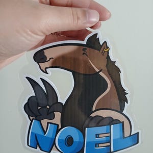 May include: A cartoon-style sticker featuring a brown and black horse-like creature with the word "NOEL" in blue block letters. The creature has large paws and a gold earring.