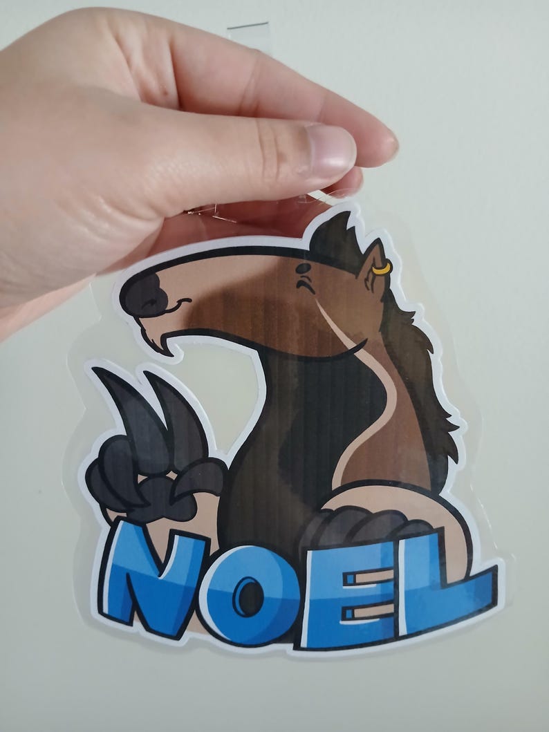 May include: A cartoon-style sticker featuring a brown and black horse-like creature with the word "NOEL" in blue block letters. The creature has large paws and a gold earring.