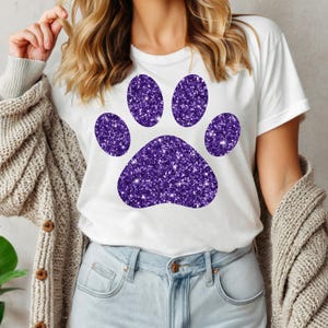 Può includere: Una maglietta bianca con un grande disegno di impronta di zampa di glitter viola.