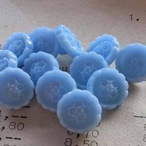 Peut inclure: Un ensemble de boutons bleus clairs en forme de fleurs. Chaque bouton a un bord festonné et un centre floral détaillé. Les boutons sont disposés sur un fond de papier vintage, créant une esthétique charmante et artisanale.