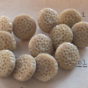 May include: A collection of twelve beige, floral-patterned buttons. Each button features a detailed, carved design of a central flower surrounded by intricate petals. The buttons are arranged on a textured, off-white surface.