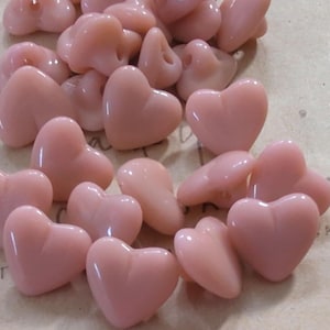 Peut inclure: Une collection de petits boutons en forme de cœur, rose clair et brillants. Chaque bouton présente une surface lisse et arrondie avec une indentation centrale, ainsi qu'une attache au dos. Les boutons sont disposés en un tas.