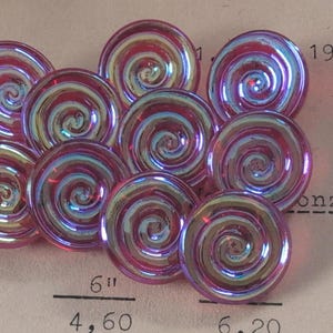 Puede incluir: Una colección de cuentas de vidrio iridiscente con remolinos. Cada cuenta tiene forma circular con un diseño en espiral en tonos rosa, morado y dorado. Las cuentas miden aproximadamente 3,8 cm de diámetro.