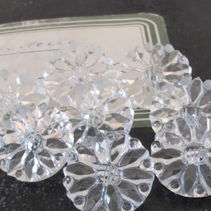 May include: Six clear, faceted crystal flower-shaped embellishments. Each has a central crystal and petal-like facets radiating outwards. The crystals are arranged on a dark surface, with a vintage label in the background.