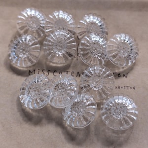 May include: A collection of eleven clear, circular glass buttons featuring a radial, sunburst-like pattern of raised ridges extending from the center to the outer edge. The buttons are arranged in a cluster on a light brown paper surface with text.