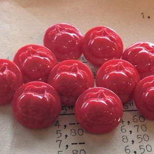 Op de afbeelding: Een verzameling van tien glanzende, ronde, rode knopen. Elke knoop heeft een verhoogd appelontwerp. De knopen liggen op een vintage papieren achtergrond met vervaagde tekst en cijfers. Diameter is ongeveer 2.5 cm.