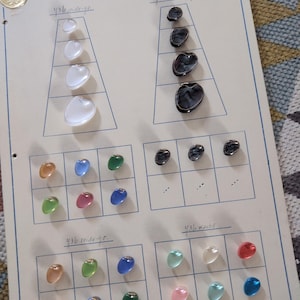 Peut inclure: Une page de catalogue vintage présentant une variété de perles de verre de différentes formes et couleurs. Les perles sont disposées en rangées et en colonnes, avec des étiquettes telles que "4/36 seide-ge" et "4/36 wachs". Les couleurs incluent le transparent, le blanc, le noir et diverses nuances de bleu, vert, rose et orange.