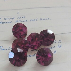 May include: Five round, dark pink faceted gemstones are displayed on lined paper. The gems have a brilliant cut, reflecting light. Handwritten text is visible in the background.