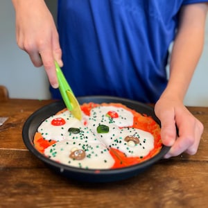 Op de afbeelding: Een kind gebruikt een groen plastic mes om een pizza te snijden gemaakt van koekdeeg, rode glazuur, witte glazuur en groene en bruine sprinkles.