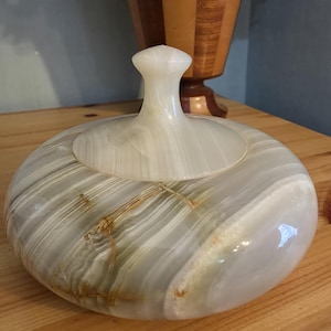 May include: A circular, lidded container crafted from polished onyx. The container features a predominantly white and grey colour scheme, with delicate green and brown veining. The lid is topped with a small, rounded handle. It rests on a wooden surface.