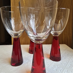 May include: Four clear wine glasses with a red base and stem. The glasses have a wide bowl and a conical stem. The glasses are arranged on a white surface.