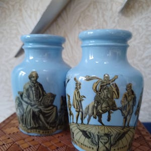 May include: Two pale blue ceramic vases with historical illustrations. One vase shows a seated figure reading a book, the other depicts a man on horseback with the text "ALL THE WORLD'S A STAGE".