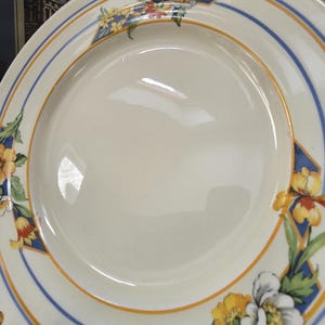 May include: Cream ceramic plate with a floral pattern. The plate has a blue and gold border with floral accents in yellow, white, and red. The flowers are arranged in geometric patterns around the rim, creating a decorative tableware piece.
