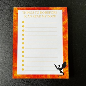 May include: A white notepad with a fire-themed border and a black silhouette of a dragon in the bottom right corner. The notepad has a title that reads "Things to do before I can read my book" and a list of bullet points.