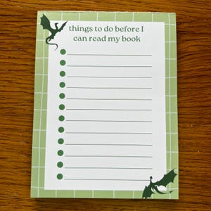 Peut inclure: Bloc-notes avec une bordure en grille verte et le texte "things to do before I can read my book". Le bloc-notes comporte une liste de lignes avec des points verts et des silhouettes de dragons dans les coins.