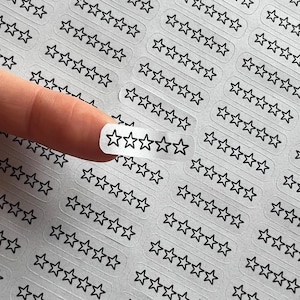 May include: Clear sticker sheet with multiple star-shaped outlines. A finger holds a sticker with five black stars. The stickers are arranged in rows and columns, creating a pattern of stars.