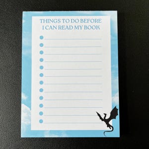 Peut inclure: Un bloc-notes bleu et blanc avec une silhouette noire de dragon dans le coin inférieur droit. Le bloc-notes porte le texte "Things to do before I can read my book" en haut.