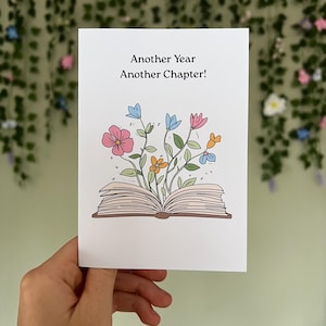 Pode incluir: Um cartão branco com o texto "Another Year Another Chapter!" apresenta uma ilustração de um livro aberto com flores e folhas coloridas brotando dele. O cartão é segurado em uma mão contra um fundo de vegetação.