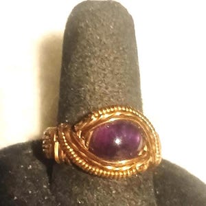Puede incluir: Anillo artesanal con una piedra preciosa púrpura engastada en un diseño de alambre dorado. El intrincado trabajo de metal rodea la piedra de forma ovalada, creando una joya única y artística.