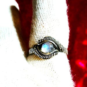 Könnte beinhalten: Ein silberfarbener Ring mit Drahtwickel-Design. Der Ring hat einen runden, irisierenden Edelstein in der Mitte. Der Edelstein hat eine blaue und weiße Farbe. Der Ring wird auf einer weißen Oberfläche vor rotem Hintergrund präsentiert.
