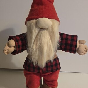 May include: A fabric gnome figure with a red pointed hat and a long white beard. The gnome is dressed in a black and red checked jacket, red trousers, and brown boots. The doll has its arms outstretched.