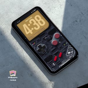 Puede incluir: Un smartphone con una funda negra y un fondo de pantalla personalizado que se asemeja a un mando de juego retro. La pantalla muestra la hora, la fecha y una interfaz temática de juego con botones rojos y una cruceta. El teléfono está sobre una superficie gris.