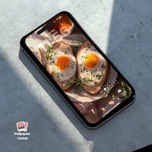 May include: A smartphone displays a wallpaper featuring two sunny-side-up eggs on toast, garnished with herbs. The screen shows the time as 11:56 and the date as October 14th. The phone is black with a silver frame, and the logo "Wallpapers Central" is visible.