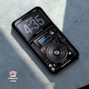 Peut inclure: Smartphone noir affichant une interface Pioneer DJ. L'écran montre une table de mixage DJ numérique avec une platine, des boutons et des curseurs. L'heure est 16h35 le mercredi 15 octobre. Le téléphone est posé sur une surface grise.