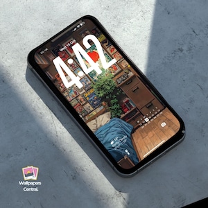 May include: A smartphone displays a wallpaper featuring a room filled with comic books, a plant, and a blue blanket. The time 4:42 is prominently displayed on the screen. The phone is black and rests on a gray surface.