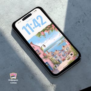 May include: A black smartphone displays a scenic wallpaper of a coastal town with pink buildings, blue water, and a light blue sky. The time is 11:42. The phone is on a gray surface. The logo for Wallpapers Central is in the bottom left corner.