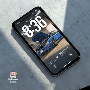 May include: A black smartphone displays a wallpaper featuring a blue sports car with its hood open, and a person working on the engine. The time 8:36 is displayed in large white numbers. The phone is on a gray surface.