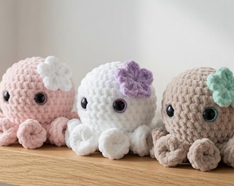 Puede incluir: Tres peluches de pulpo tejidos a ganchillo con hilo suave. El de la izquierda es rosa claro con una flor blanca, el del medio es blanco con una flor morada y el de la derecha es marrón claro con una flor verde menta. Cada uno tiene ojos negros grandes.