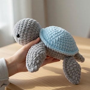 May include: A plush turtle toy crafted from soft, chunky gray yarn with a light blue shell. The turtle features a rounded head, a textured crocheted surface, and a small black bead eye. The design is simple, focusing on the soft, tactile material.
