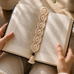 May include: A cream-coloured crochet bookmark featuring a series of connected circular floral motifs. The bookmark is placed inside an open book and is finished with a single dark brown wooden bead and a small matching cream-coloured yarn tassel.