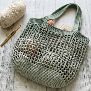 May include: A sage green crocheted tote bag featuring an open mesh body and solid base with two integrated handles. A ball of beige yarn and a gold-toned crochet hook are positioned nearby, highlighting the handmade nature of the textile accessory.