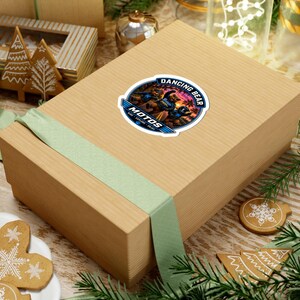 Puede incluir: Una pegatina circular con el gráfico de dos osos en motocicleta frente a un atardecer. El texto dice DANCING BEAR MOTOS RIDE EXPLORE ENJOY. La pegatina está colocada sobre una caja de regalo rectangular de color marrón claro con cinta verde.