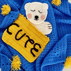 May include: A blue crocheted baby blanket with a white crocheted polar bear applique and a yellow crocheted patch that says "cute".