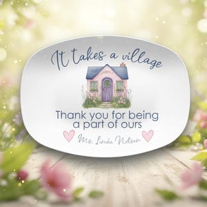 Pode incluir: Prato oval de cerâmica branca com uma ilustração em aquarela de uma casa rosa com telhado azul. O texto diz "It takes a village" e "Thank you for being a part of ours". Dois corações rosa estão em baixo.