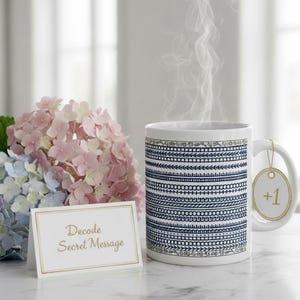 May include: A white ceramic mug with a blue and white pattern, steam rising from it. A small tag with "+1" hangs from the handle. A card with "Decode Secret Message" is in front of a bouquet of pink and blue hydrangeas.