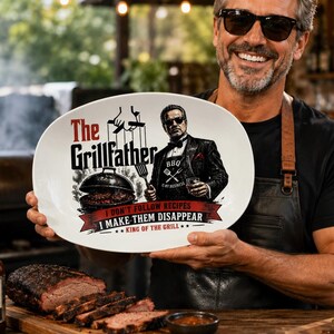 Op de afbeelding: Een ovale serveerschaal met de tekst "The Grillfather" en een afbeelding van een man in een pak die een vork en een drankje vasthoudt. De schaal heeft ook de tekst "I don't follow recipes, I make them disappear." Een fles BBQ-saus en een hamburger zijn ook zichtbaar.