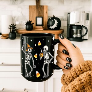 以下が含まれることがあります： 黒いセラミックハロウィーンマグカップ。踊る骸骨、キャンディーコーン、星が描かれています。骸骨の一人は魔女の帽子をかぶり、手を振っています。もう一人の骸骨はマグカップを持っています。マグカップの取っ手は黒です。