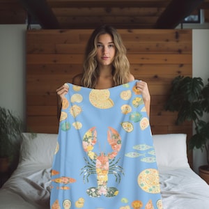 May include: A light blue blanket featuring a large lobster illustration filled with floral patterns. The blanket also includes illustrations of seashells and fish in shades of yellow, orange, and green. The blanket is held up in front of a bed.