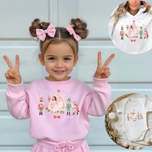 May include: A young girl wearing a pink sweatshirt and skirt, with a matching bow in her hair, makes a peace sign. The sweatshirt features a Christmas-themed graphic with a tree, nutcrackers, and other festive characters. A white hoodie and onesie with the same graphic are also shown.