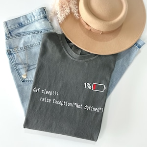Peut inclure: Composition à plat avec un t-shirt gris anthracite portant l'inscription "def sleep(): raise Exception("Not defined")" et une icône de batterie à 1%. Le t-shirt est associé à un jean bleu clair et un chapeau fedora beige.