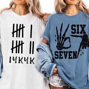 May include: Two t-shirts, one white and one blue, with black graphic designs. The white shirt has tally marks and the text "14KUK". The blue shirt features a skeleton hand gesture and the words "SIX SEVEN".