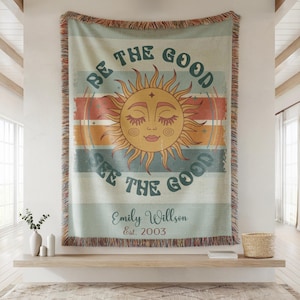 May include: A woven throw blanket with a sun design and the text "BE THE GOOD SEE THE GOOD". The blanket has a fringed edge and includes the name "Emily Willson" and the year "Est. 2003". The color palette includes shades of blue, orange, and cream.