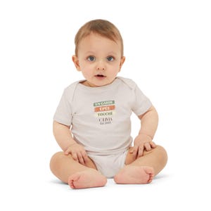 May include: A baby wearing a light beige onesie with the words "EN GARDE ÉPÉE TOUCHÉ OLIVIA Est. 2023" printed in green, orange, and brown. The baby is sitting with legs crossed and looking at the camera.